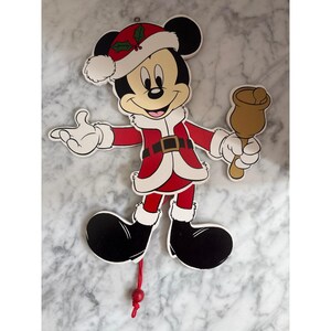 May include: A flat, die-cut wall decoration of Mickey Mouse dressed in a red and white Santa Claus suit with a matching hat featuring holly leaves. The character holds a brown hand bell in one hand and has a red pull string with a bead at the bottom.