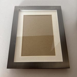 May include: A rectangular picture frame with a brushed metal finish. The frame has a white mat and a brown backing. The frame is designed to hold a photograph or artwork. The frame is a neutral color and can be used in any room.