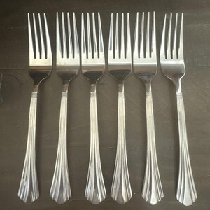 May include: Six stainless steel forks arranged in a row. Each fork has a long, slender handle with a decorative, fan-like design at the base. The tines are evenly spaced and the forks are set against a dark background.
