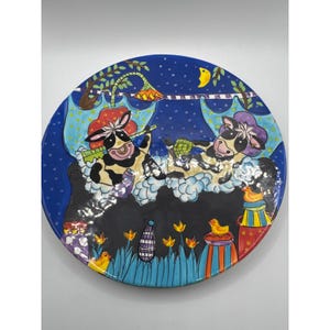 May include: A round ceramic plate featuring a dark blue night sky with a yellow crescent moon. Two black and white cows are depicted soaking in a bubble bath under a light blue umbrella. The foreground shows orange tulips and yellow ducks.