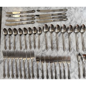 May include: A collection of vintage silverware, including knives, forks, and spoons, arranged on a marble surface. The silverware has ornate floral detailing on the handles. The set includes various sizes of spoons and forks.
