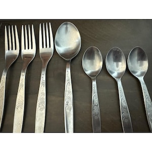 May include: A set of stainless steel silverware, including forks and spoons. The handles feature a floral design. The spoons vary in size, with a large serving spoon and smaller teaspoons. The silverware is arranged on a dark surface.