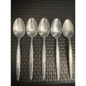 May include: Five silver spoons arranged in a row. Each spoon has a rounded bowl and a handle with a decorative, etched pattern. The spoons are made of a shiny, reflective metal, likely stainless steel, and are set against a dark background.