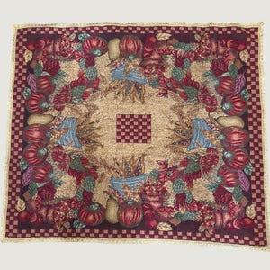 May include: A square woven tapestry table topper featuring a central checkered square surrounded by a harvest-themed pattern of cornucopias, red pumpkins, and green foliage. The border consists of a red and tan checkered pattern with fringe.