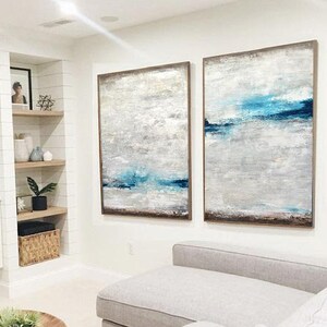 Puede incluir: Dos pinturas abstractas en un sal&oacute;n. Las pinturas son de color azul claro, blanco y gris con un marco marr&oacute;n. Las pinturas est&aacute;n colgadas en una pared blanca sobre un sof&aacute; blanco.