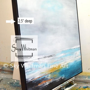 May include: A large abstract painting on canvas with a black frame. The painting depicts a seascape with shades of blue, white, and gray. The frame is 2.5 inches deep. The painting is signed "Sky Whitman Original Modern Art" in the lower left corner.