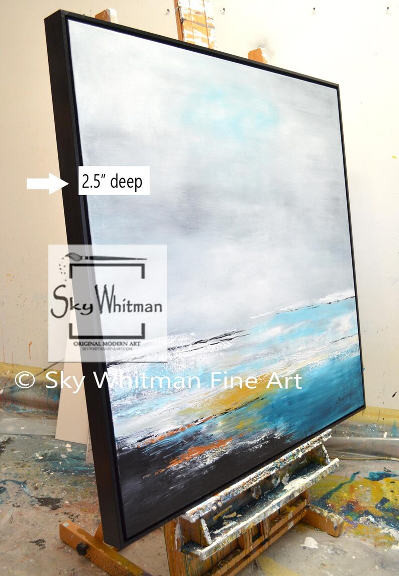 May include: A large abstract painting on canvas with a black frame. The painting depicts a seascape with shades of blue, white, and gray. The frame is 2.5 inches deep. The painting is signed "Sky Whitman Original Modern Art" in the lower left corner.