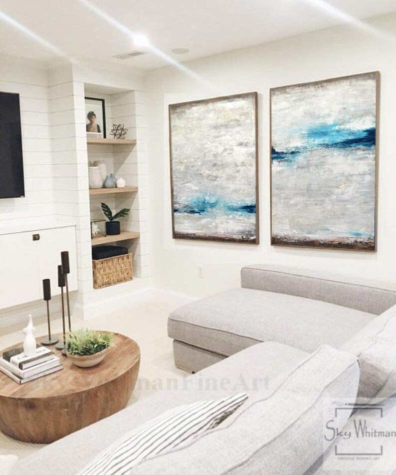 Puede incluir: Dos pinturas abstractas en un sal&oacute;n. Las pinturas son de color azul claro, blanco y gris con un marco marr&oacute;n. Las pinturas est&aacute;n colgadas en una pared blanca sobre un sof&aacute; blanco.