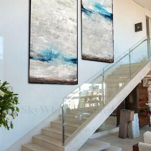 Puede incluir: Dos pinturas abstractas con un esquema de color blanco, gris y azul cuelgan de una pared blanca sobre una escalera moderna con barandillas de vidrio.