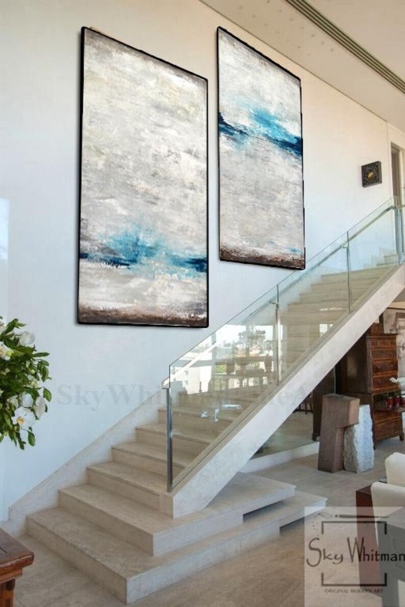 Puede incluir: Dos pinturas abstractas con un esquema de color blanco, gris y azul cuelgan de una pared blanca sobre una escalera moderna con barandillas de vidrio.