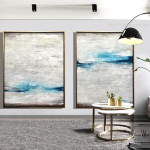 Puede incluir: Dos pinturas abstractas en una sala de estar moderna. Las pinturas son en tonos de azul, blanco y gris. Las pinturas est&aacute;n enmarcadas en oro y colgadas en una pared blanca. Las pinturas est&aacute;n sobre un conjunto de dos mesas redondas anidadas con patas de metal dorado.