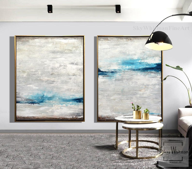 Puede incluir: Dos pinturas abstractas en una sala de estar moderna. Las pinturas son en tonos de azul, blanco y gris. Las pinturas est&aacute;n enmarcadas en oro y colgadas en una pared blanca. Las pinturas est&aacute;n sobre un conjunto de dos mesas redondas anidadas con patas de metal dorado.