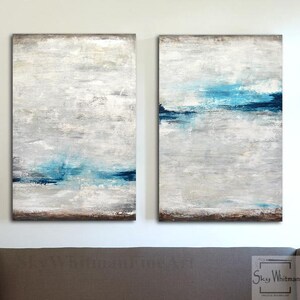 Puede incluir: Un conjunto de dos pinturas abstractas con fondo blanco y pinceladas azules y grises. Las pinturas est&aacute;n colgadas en una pared blanca sobre un sof&aacute; marr&oacute;n.