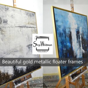May include: Two abstract paintings in gold metallic floater frames. The painting on the left is white, gray, and black. The painting on the right is blue, white, and black. Both paintings are on easels.
