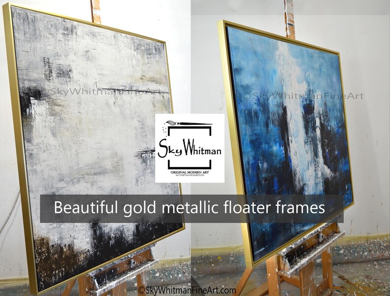May include: Two abstract paintings in gold metallic floater frames. The painting on the left is white, gray, and black. The painting on the right is blue, white, and black. Both paintings are on easels.