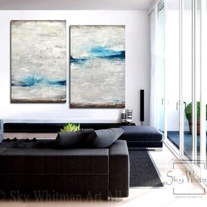 Puede incluir: Dos pinturas abstractas en una sala de estar moderna. Las pinturas son de color gris claro con l&iacute;neas horizontales azules. Las pinturas est&aacute;n colgadas en una pared blanca sobre un sof&aacute; negro.