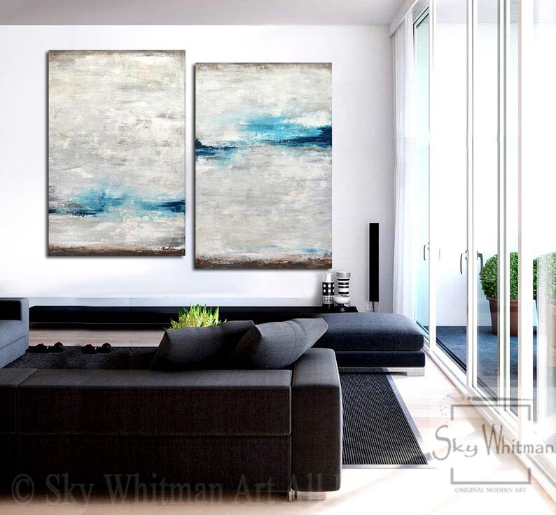 Puede incluir: Dos pinturas abstractas en una sala de estar moderna. Las pinturas son de color gris claro con l&iacute;neas horizontales azules. Las pinturas est&aacute;n colgadas en una pared blanca sobre un sof&aacute; negro.
