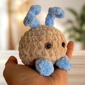 May include: A handmade plush toy in the shape of a round creature, held in a hand. The body is a light brown color, with blue accents on the feet and antennae. The toy has two dark eyes.