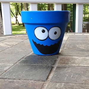 Op de afbeelding: Een felblauwe terracotta plantenpot met een handgeschilderd gezicht met twee grote witte ronde ogen, zwarte pupillen en een brede, grillige zwarte mond. De pot heeft een uitlopende rand en een taps toelopende basis met een gladde afwerking.