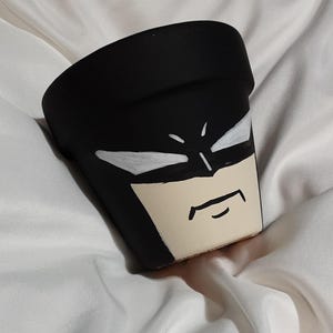 Puede incluir: Una maceta de terracota negra con una ilustración pintada a mano de la cara de Batman. La maceta tiene un fondo blanco y presenta la máscara icónica de Batman con una expresión seria.