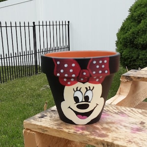 May include: Black painted flower pot featuring a smiling cartoon face with a red bow with white polka dots. The interior of the pot is orange. The pot is sitting on a wooden surface.