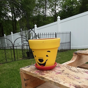 May include: A yellow painted flower pot with a smiling cartoon face, sitting on a wooden surface. The pot has a red saucer and is set against a backdrop of a white fence and green grass. The pot is approximately 7 inches tall.