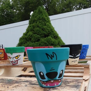以下が含まれることがあります： キャラクターデザインが施された、塗装されたセラミック植木鉢のコレクション。鉢は、スティッチ、バットマン、ジョーカーなど、さまざまな色とデザインで塗装されています。鉢は木製のパレットに展示されています。