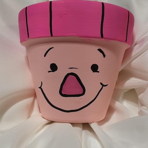 以下が含まれることがあります： 手描きのテラコッタ植木鉢で、笑顔のデザインです。 植木鉢は、黒い目、鼻、笑顔の口が付いたピンク色のベースが特徴です。 リムは黒い縦縞模様のピンク色に塗られています。 この植木鉢は植物を入れるように設計されています。