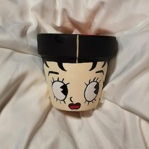 Peut inclure: Un pot de fleurs en terre cuite peint à la main avec un visage de femme de dessin animé aux cheveux noirs, de grands yeux et des lèvres rouges. Le pot est peint en noir, blanc, jaune et rouge.