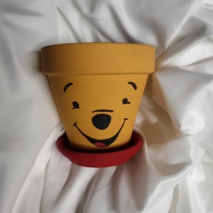 Puede incluir: Una maceta de cerámica amarilla con una cara de Winnie the Pooh pintada. La maceta tiene un borde rojo y un platillo rojo.