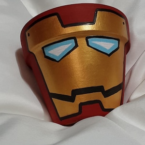 Puede incluir: Una maceta de cerámica roja y dorada con un diseño pintado de la cara de Iron Man. La maceta tiene un contorno negro y ojos azules.