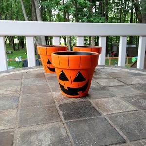 May include: Three orange terracotta pots painted with black jack-o'-lantern faces. Each pot features a wide rim, a tapered body, and vertical black lines mimicking pumpkin ridges. The interior of each pot is painted black, contrasting with the bright orange exterior.