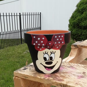 May include: A terracotta flower pot painted black with a cream-colored cartoon character face featuring large eyes, long eyelashes, and a wide smile. A red bow with white polka dots is painted across the top rim of the pot, which has an orange interior.