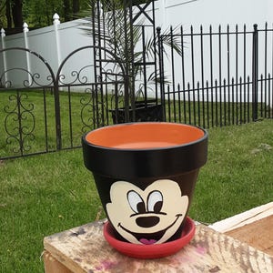 Puede incluir: Una maceta pintada de negro y naranja con un platillo rojo. La maceta presenta una cara de Mickey Mouse pintada. La maceta está sobre una superficie de madera, con un césped verde y una valla blanca en el fondo.