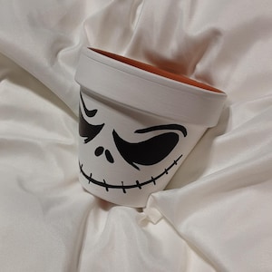 Peut inclure: Pot en céramique blanche avec un visage de Jack Skellington peint en noir. Le pot a un bord en terre cuite brune.