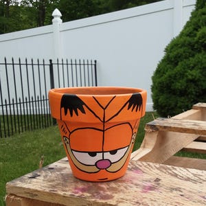 May include: Orange ceramic plant pot with a hand-painted Garfield cartoon character design. The pot is round and sits on a wooden surface. The Garfield face features black, white, pink, and orange colours.