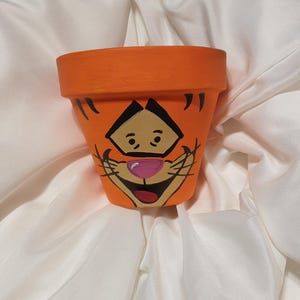 Op de afbeelding: Oranje terracotta bloempot met een handgeschilderd cartoon gezicht van een tijger. De tijger heeft zwarte ogen, een roze neus en een brede glimlach.