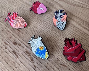 May include: A collection of heart-shaped enamel pins in various designs. One pin features the text "LOVE HURTS", while others depict anatomical details, hands, and abstract patterns. The pins are in shades of red, pink, white, blue, and yellow.