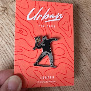 Puede incluir: Un pin de esmalte metálico que muestra una figura negra y plateada lanzando un ramo de flores amarillas, montado sobre una tarjeta de soporte rectangular naranja con el texto Urban PIN CLUB LONDON e IG: tomadoorshop en blanco.