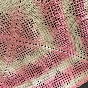 May include: A handmade crochet blanket in shades of pink and white. The blanket features a square pattern with openwork detailing. The color gradient transitions from white to pink, creating a soft, textured appearance. The blanket is likely made of soft yarn.