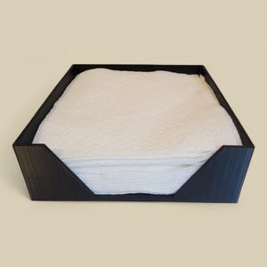 May include: A black square napkin holder with a slanted front edge, filled with a stack of white paper napkins. The holder is made of a solid material and sits on a neutral background.