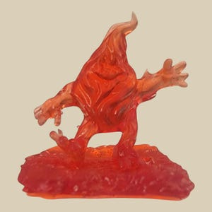 May include: A translucent orange figurine of a stylized creature with a flame-like body and outstretched arms. The figure stands on a textured, reddish-orange base, with a light beige background.