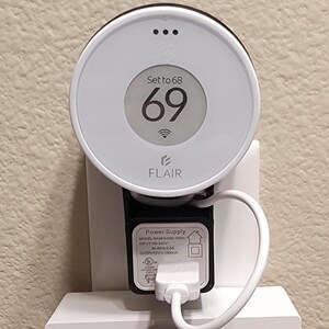 May include: A white smart thermostat with a digital display showing a temperature of 69 degrees. The thermostat is labeled "FLAIR" and has a power supply cord plugged in. The text on the power supply reads "Power Supply, MODEL SAWOS-050-1000, INPUT 100-240V 50-60Hz 0.3A, OUTPUT 5V 1000mA."