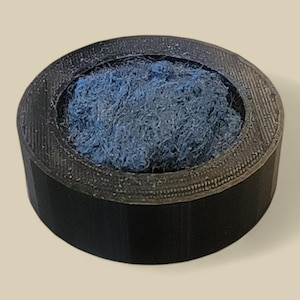 May include: A black, cylindrical object with a textured rim, filled with a mass of blue fibrous material. The object appears to be made of a hard, possibly 3D-printed material. The blue material contrasts with the black exterior.