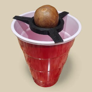 Può includere: Un bicchiere di plastica rosso con bordo bianco contiene un oggetto marrone, forse un seme o una noce, sostenuto da un supporto nero a forma di stella. Il bicchiere è posto su uno sfondo neutro.
