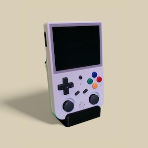 Op de afbeelding: Een witte handheld gameconsole met een zwart scherm, vier gekleurde knoppen en twee zwarte joystickknoppen. De console staat op een zwarte standaard.