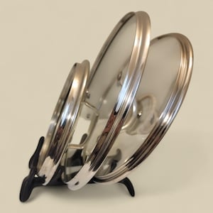 May include: Three stainless steel and glass pot lids in a black metal holder. The lids have shiny rims and clear glass centres. The holder is designed to hold the lids upright. The holder is black.