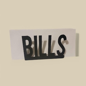 May include: Black metal sign with the word "BILLS" in all caps. The sign is sitting on a white piece of paper.