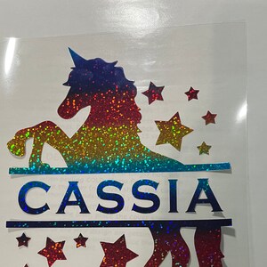 May include: A rainbow glitter unicorn silhouette with the name "CASSIA" in blue glitter letters. The unicorn is surrounded by small red glitter stars.