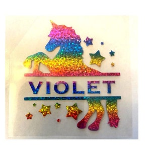 May include: A colorful unicorn design with the name "VIOLET" in blue letters. The unicorn and stars are in a rainbow glitter pattern. The design is on a clear background.